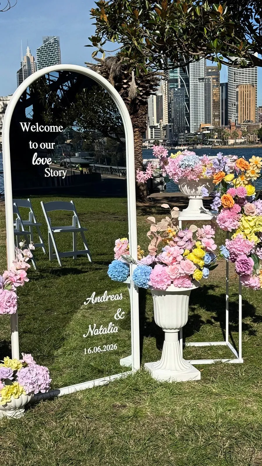 Wedding welcome mirror reading “Welcome to our love story” framed by pastel florals, with the city skyline behind