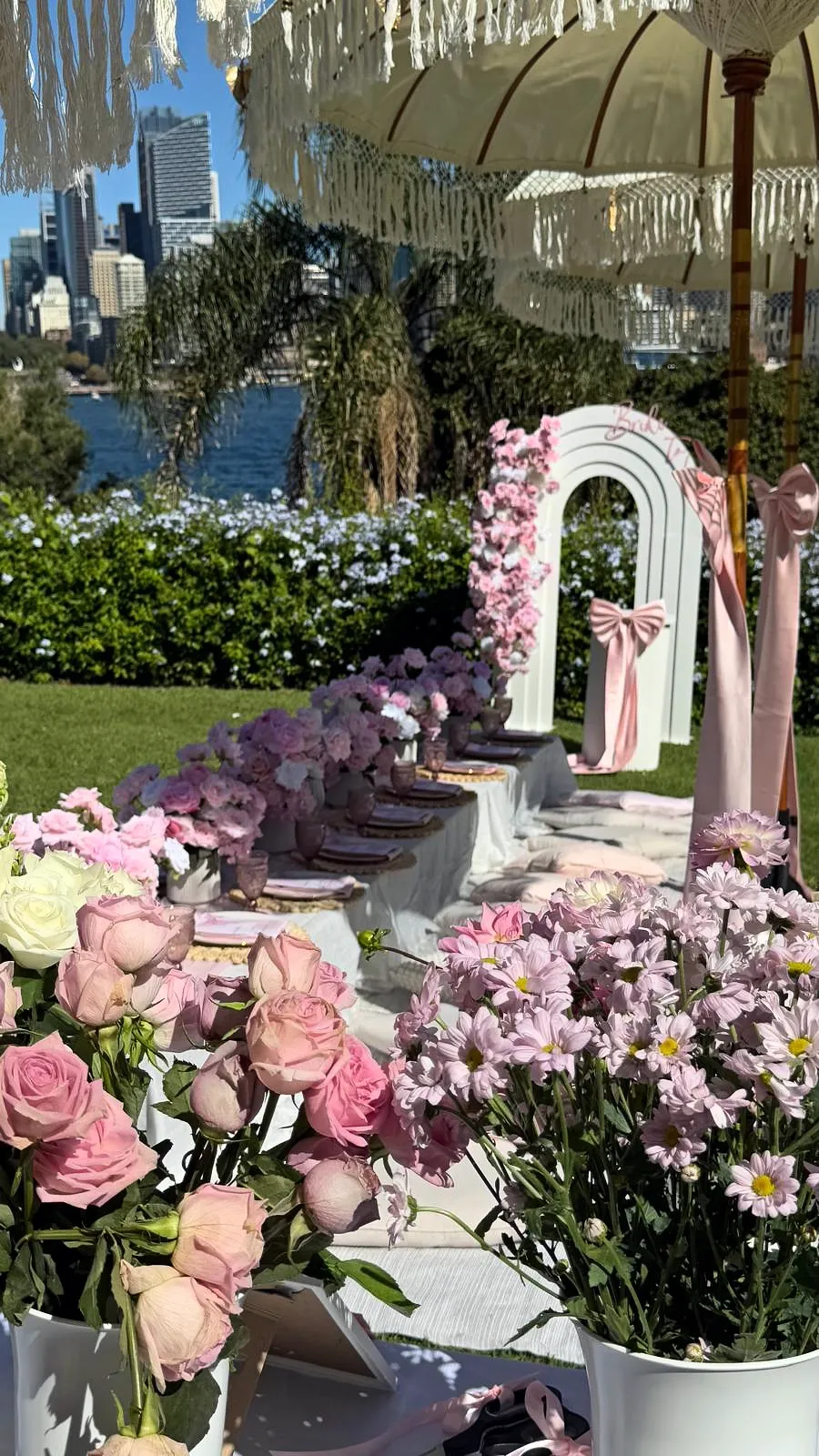 Pink luxury picnic with a rose-covered arch, low tables, cushions and fringed umbrellas, with the Sydney city skyline behind
