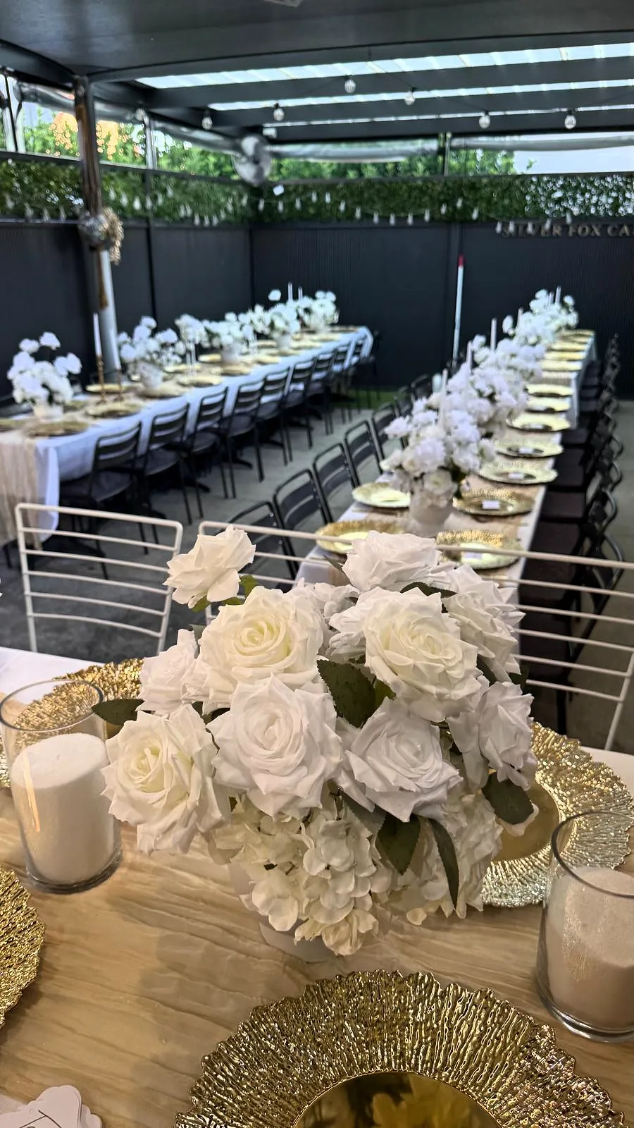 Long tables with white rose centrepieces, gold chargers and candles under a pergola